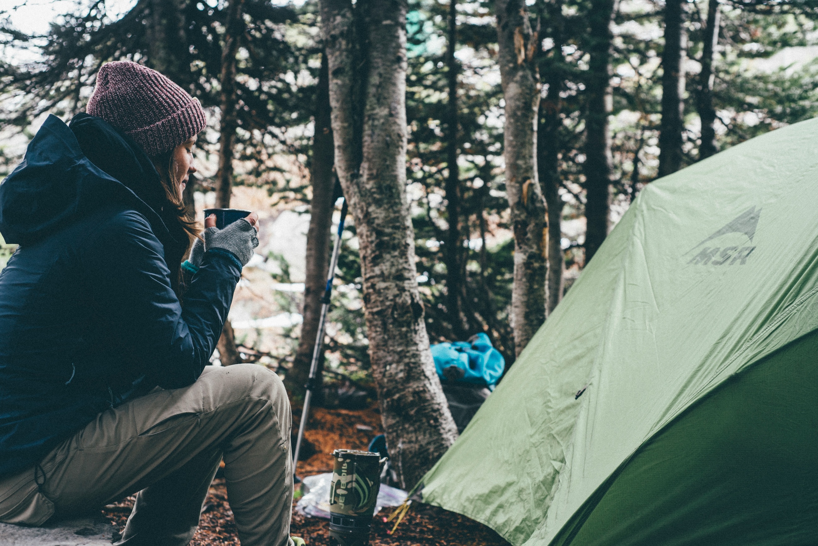 A camper sitting with a hot drink beside a tent at a wooded campsite