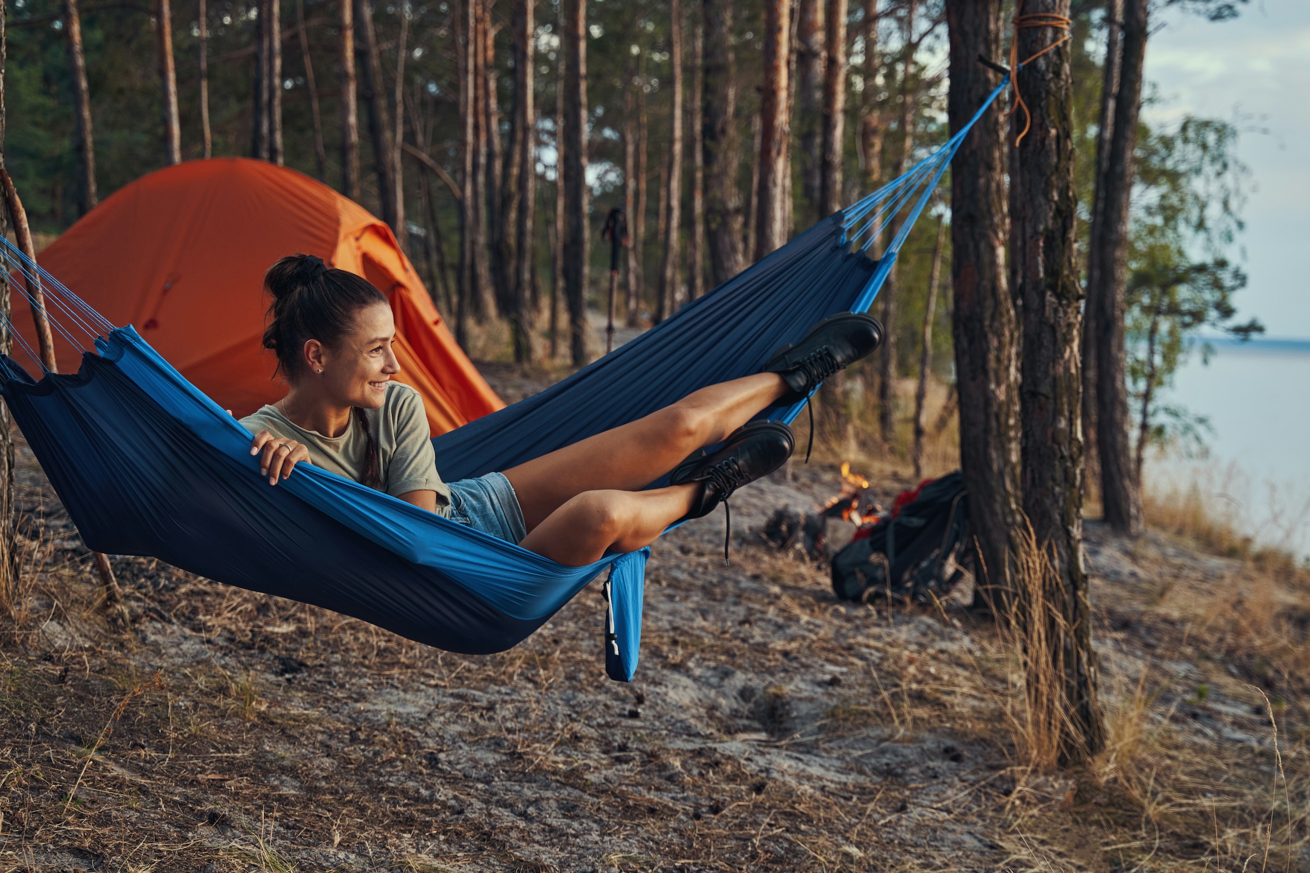 A camper resting in a hammock beside a tent and a small fire at a lakeside campsite