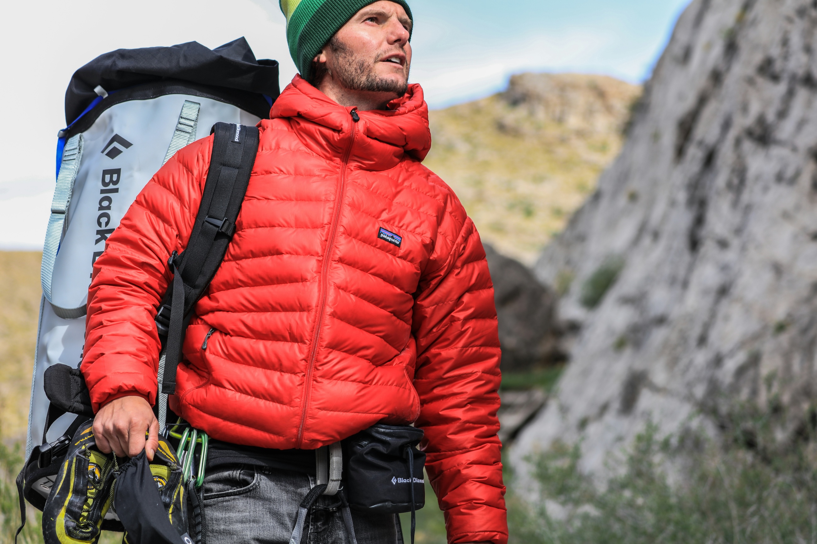 A climber in a down jacket carrying a pack and climbing shoes below a rock face