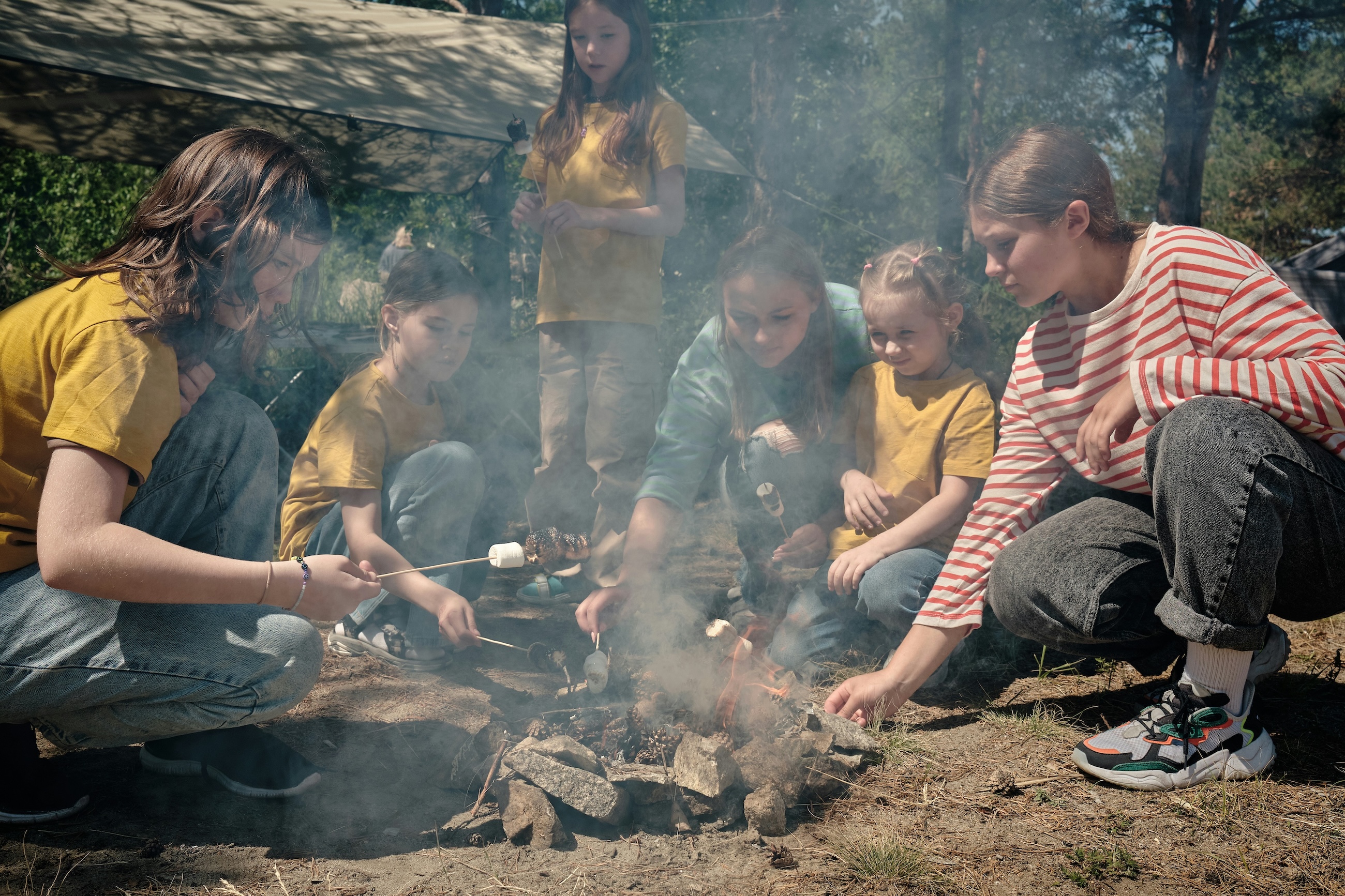 Campers gathered around a small fire under a tarp at a wooded camp site
